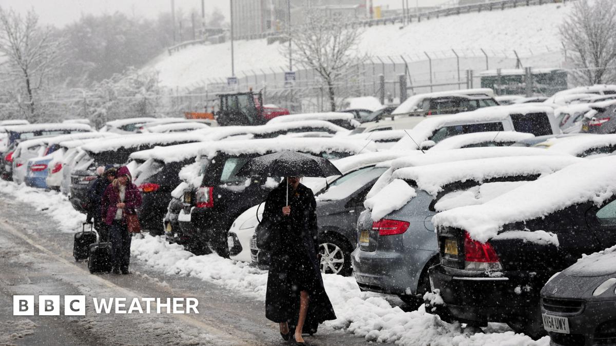 Woman walking through a car park in heavy snow carrying an umbrella